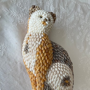 May include: A cat figurine made entirely of seashells. The cat is sitting with its tail curled around its body. The shells are arranged in a pattern of white, brown, and tan.