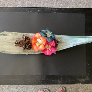 May include: A white wooden boat-shaped dish with a green and blue glaze. The dish is filled with three artificial flowers in shades of pink, orange, and blue. There are also three pine cones in the dish.