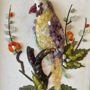 May include: A colorful mosaic bird perched on a branch with green leaves. The bird is made of small pieces of glass and stone in shades of yellow, purple, red, and green.