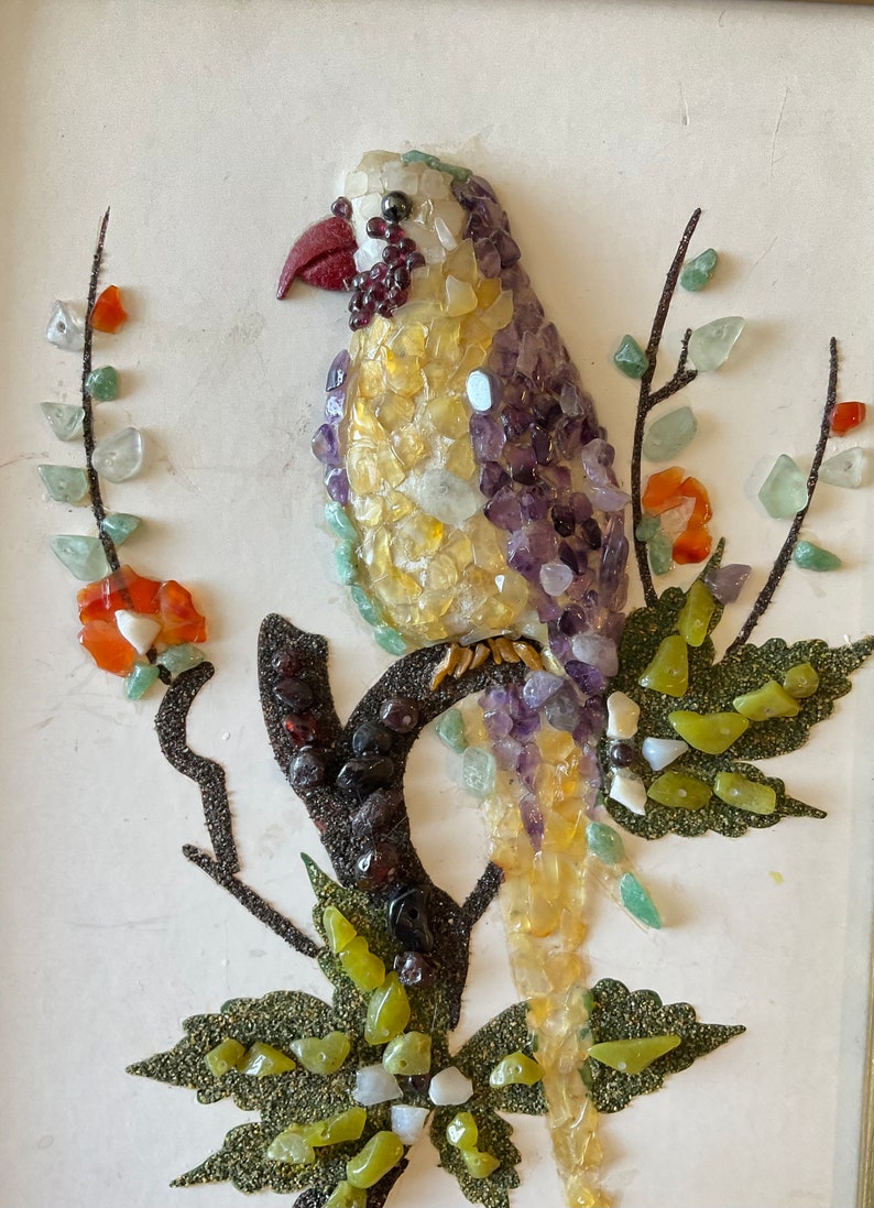 May include: A colorful mosaic bird perched on a branch with green leaves. The bird is made of small pieces of glass and stone in shades of yellow, purple, red, and green.