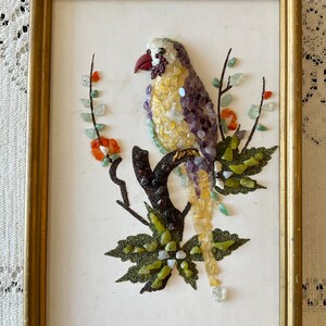 May include: A framed artwork featuring a colorful bird made of small stones and gems. The bird is perched on a branch with green leaves and red flowers. The artwork is set against a white background.