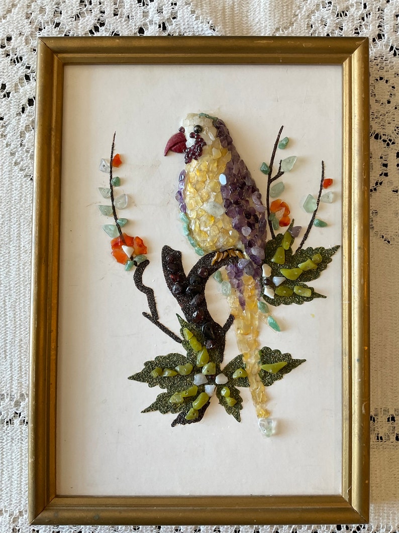 May include: A framed artwork featuring a colorful bird made of small stones and gems. The bird is perched on a branch with green leaves and red flowers. The artwork is set against a white background.