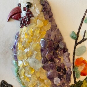 May include: A colorful mosaic bird made of small, polished stones. The bird is mostly yellow and purple, with a red beak and black eyes. The bird is perched on a branch made of small, black stones.