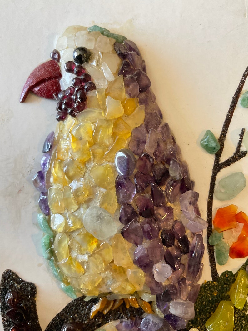 May include: A colorful mosaic bird made of small, polished stones. The bird is mostly yellow and purple, with a red beak and black eyes. The bird is perched on a branch made of small, black stones.