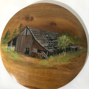 May include: A hand-painted wooden round plaque with a rustic barn scene. The barn is painted in shades of brown and gray with a weathered roof. The scene is set against a backdrop of green trees and grass. The plaque is signed "R. Staley" in the lower right corner.