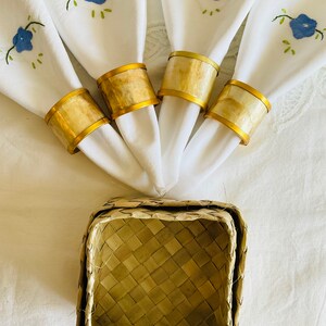 May include: Four white linen napkins with blue embroidered flowers are wrapped with gold napkin rings. The rings are made of woven natural material. A woven basket sits below the napkins.
