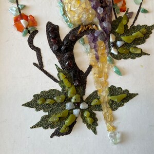 May include: A framed artwork featuring a bird made of colorful stones and gems. The bird is perched on a branch with green leaves. The artwork is set against a white background.