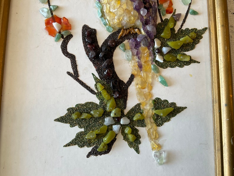 May include: A framed artwork featuring a bird made of colorful stones and gems. The bird is perched on a branch with green leaves. The artwork is set against a white background.