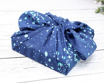 Puede incluir: Un envoltorio de regalo de tela azul con fondo blanco y un patrón de puntos azul verdoso. La tela está atada con un nudo y las palabras "Kindly Gather" están impresas en la tela.