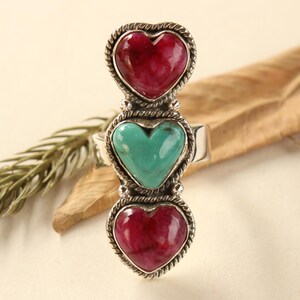 May include: A silver ring with three heart-shaped gemstones. The top and bottom hearts are red, the middle one turquoise. Each heart is framed with a rope-style border. The ring is a statement piece.
