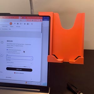 Puede incluir: Un soporte naranja para portátil está colocado junto a un portátil que muestra un anuncio de Etsy para una bandeja de servicio artesanal. El soporte es de plástico y tiene una forma única. La pantalla del portátil muestra el precio de 50,00 $.