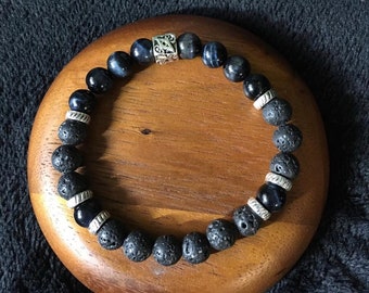 May include: A beaded bracelet featuring a mix of dark blue tiger eye, black lava rock, and silver-toned metal beads. The bracelet is displayed on a wooden surface, showcasing its natural textures and colors.
