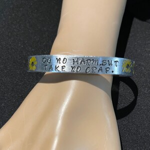May include: Silver bracelet with the text "Do no harm, but take no crap." and yellow flower accents.