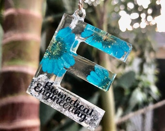 May include: A clear resin keychain in the shape of the letter E, featuring embedded bright blue daisy-like flowers and silver glitter. The bottom horizontal bar of the letter contains the black text Biomedical Engineering in a cursive script font.
