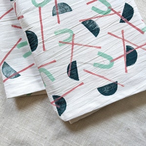 May include: Two white cotton tea towels with a geometric pattern of pink and green lines and dark blue semi-circles.