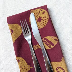 May include: A burgundy fabric napkin with a gold Christmas ornament design. A silver fork and knife are placed on top of the napkin.