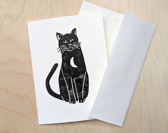 Op de afbeelding: Een zwart-witte illustratie van een kat met een halve maan op zijn borst. De kat zit met zijn staart om zijn poten gewikkeld.