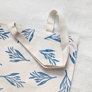 May include: A white canvas tote bag with blue floral print and cream colored straps. The bag is folded in half and the straps are visible.