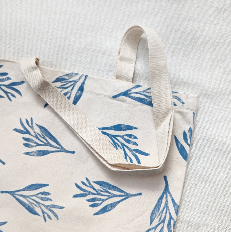 May include: A white canvas tote bag with blue floral print and cream colored straps. The bag is folded in half and the straps are visible.