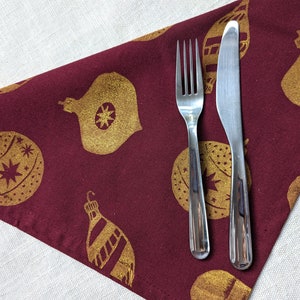 May include: A burgundy fabric napkin with a gold Christmas ornament pattern and a silver fork and knife placed on top.