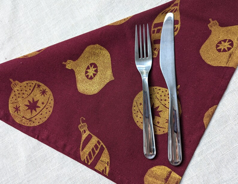May include: A burgundy fabric napkin with a gold Christmas ornament pattern and a silver fork and knife placed on top.