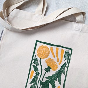 Peut inclure: Un sac fourre-tout en toile blanche avec un imprim&eacute; de pissenlits vert et jaune. Le sac porte une &eacute;tiquette indiquant "coton biologique".