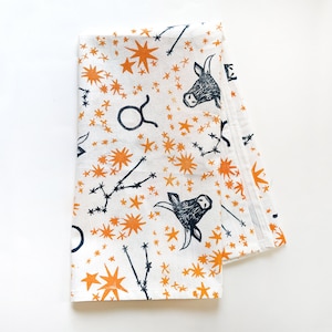 Puede incluir: Una toalla de cocina blanca con un estampado azul marino y naranja de la constelación de Tauro y una cabeza de toro. El estampado se repite en toda la toalla.