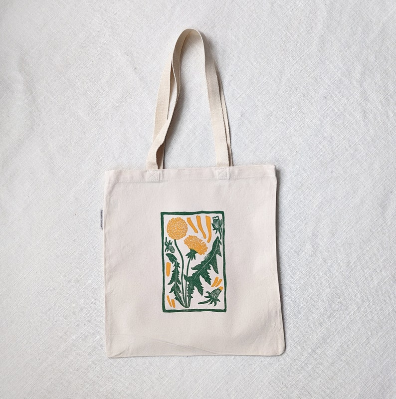 Peut inclure: Un sac fourre-tout blanc avec un imprim&eacute; de pissenlit vert et jaune. Le sac a une longue anse et est fait de tissu naturel.