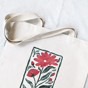 Pu&ograve; includere: Una borsa tote in tela bianca con una stampa floreale verde e rossa. La stampa presenta un grande fiore rosso con foglie verdi e fiori rossi pi&ugrave; piccoli.