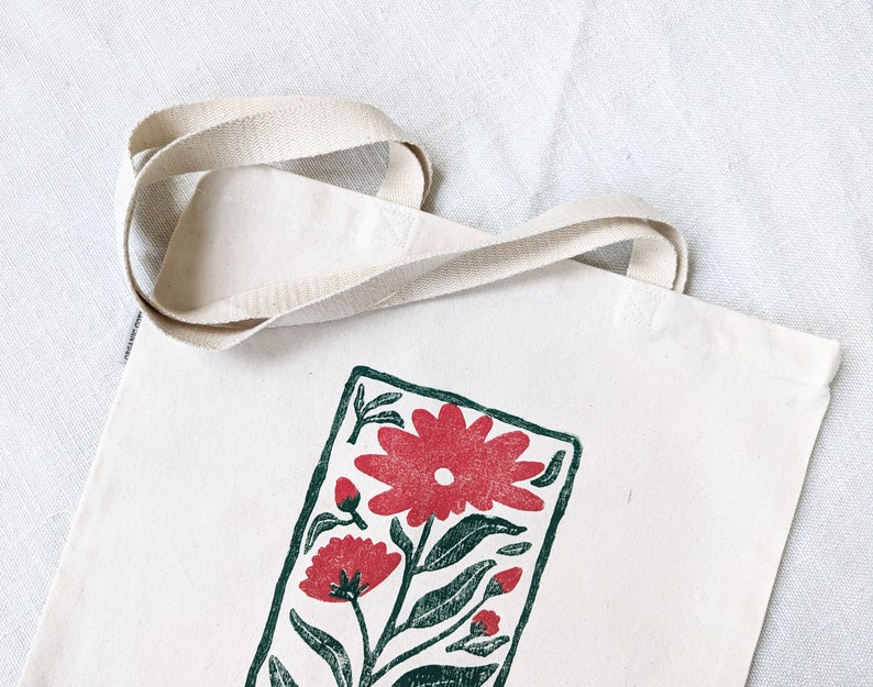 Pu&ograve; includere: Una borsa tote in tela bianca con una stampa floreale verde e rossa. La stampa presenta un grande fiore rosso con foglie verdi e fiori rossi pi&ugrave; piccoli.