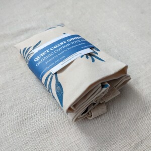 May include: A folded organic cotton tote bag with a blue and white leaf print. The bag is labeled "Quiet Coast Goods Organic Cotton Tote Bag" and includes the text "printed by hand in Portland, Maine" and the website address "quietcoastgoods.etsy.com".