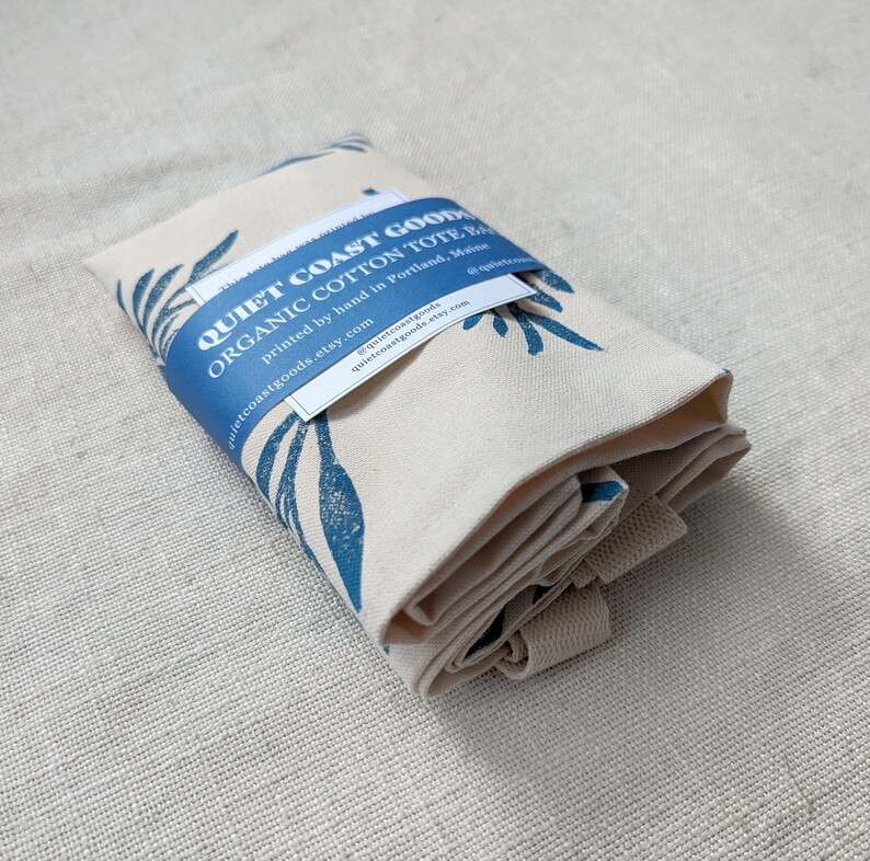 May include: A folded organic cotton tote bag with a blue and white leaf print. The bag is labeled "Quiet Coast Goods Organic Cotton Tote Bag" and includes the text "printed by hand in Portland, Maine" and the website address "quietcoastgoods.etsy.com".