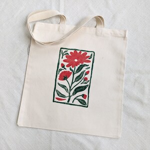 Pu&ograve; includere: Una borsa tote in tela bianca con una stampa floreale rossa e verde. La stampa &egrave; in una cornice rettangolare e presenta un grande fiore rosso con fiori rossi pi&ugrave; piccoli e foglie verdi.