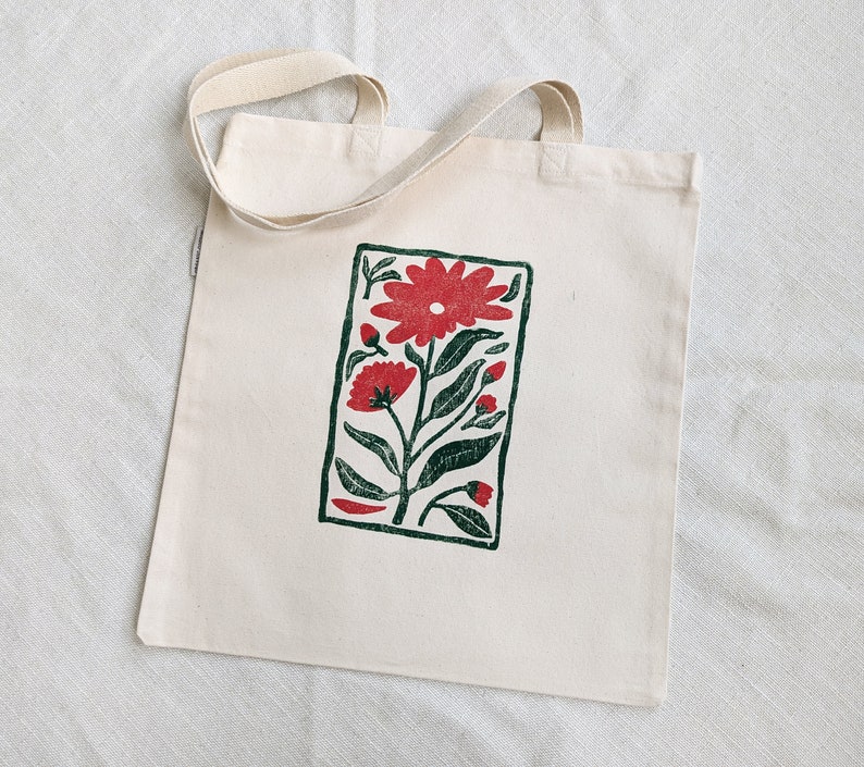 Pu&ograve; includere: Una borsa tote in tela bianca con una stampa floreale rossa e verde. La stampa &egrave; in una cornice rettangolare e presenta un grande fiore rosso con fiori rossi pi&ugrave; piccoli e foglie verdi.