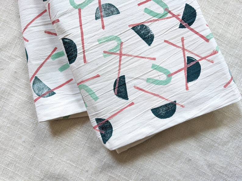 May include: Two white cotton tea towels with a geometric pattern of pink and green lines and dark blue semi-circles.
