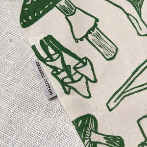 May include: A close-up of a white tote bag with a green mushroom print. The bag has a label that says "Organic Cotton".