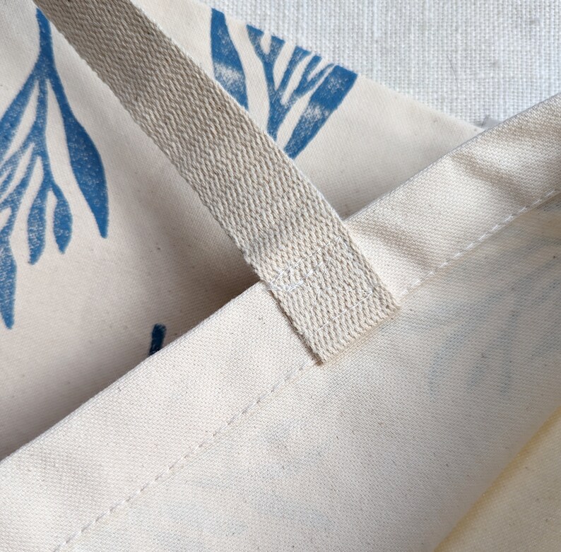 May include: Close-up of a cream-colored tote bag with blue floral print. The bag has a thick, cream-colored cotton strap.