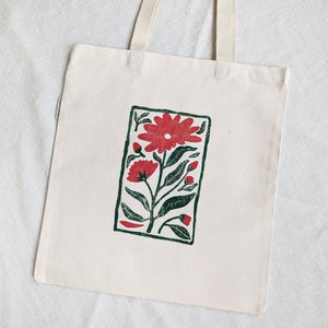 Pu&ograve; includere: Una borsa tote in tela bianca con una stampa floreale rossa e verde. La stampa presenta un grande fiore rosso con foglie e steli verdi.