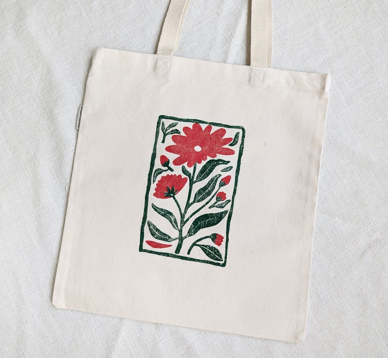 Pu&ograve; includere: Una borsa tote in tela bianca con una stampa floreale rossa e verde. La stampa presenta un grande fiore rosso con foglie e steli verdi.