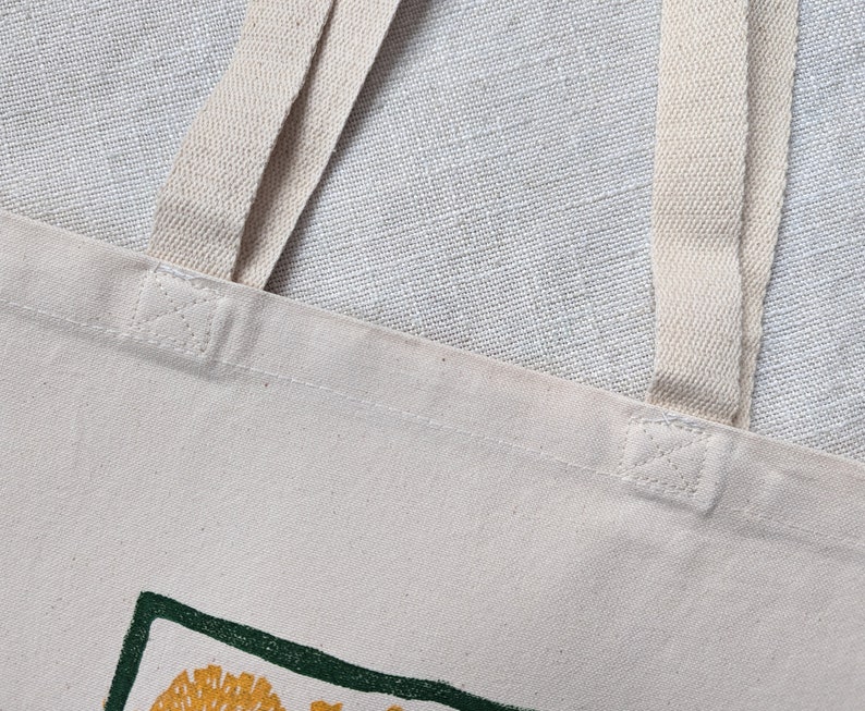 Peut inclure: Gros plan sur un sac fourre-tout en toile blanche avec deux anses cr&egrave;me. Le sac a un motif vert et jaune sur le devant.