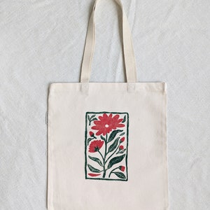 Pu&ograve; includere: Una borsa tote in tela bianca con un bordo verde e un motivo floreale rosso stampato sul davanti.