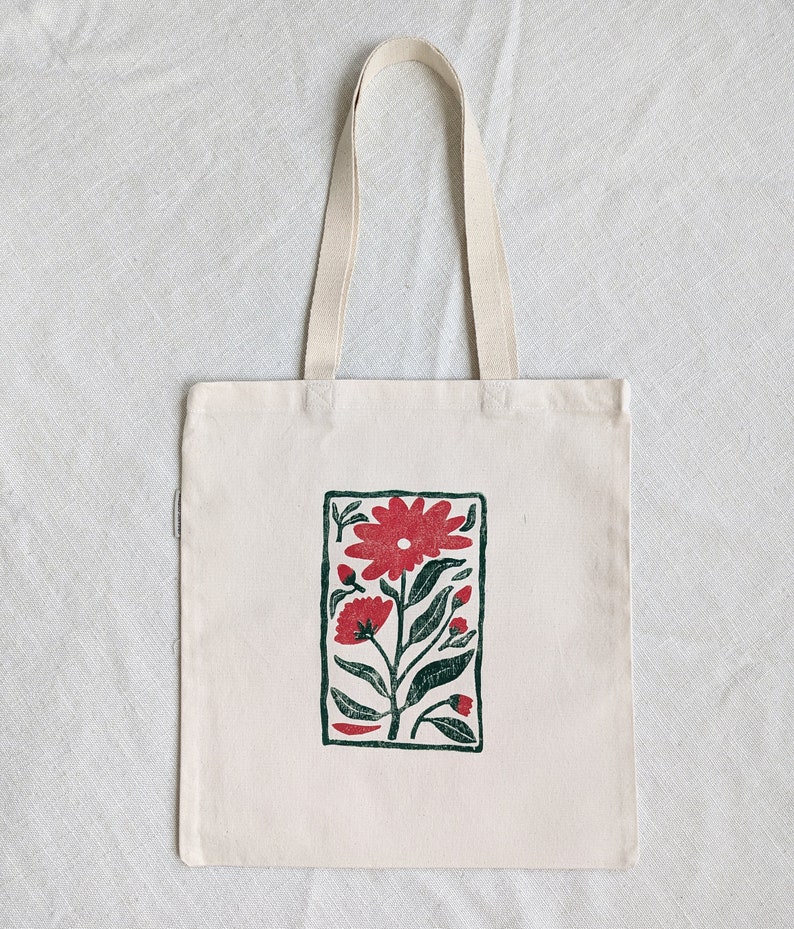 Pu&ograve; includere: Una borsa tote in tela bianca con un bordo verde e un motivo floreale rosso stampato sul davanti.
