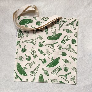 May include: A white canvas tote bag with green mushroom illustrations. The bag has two long straps.
