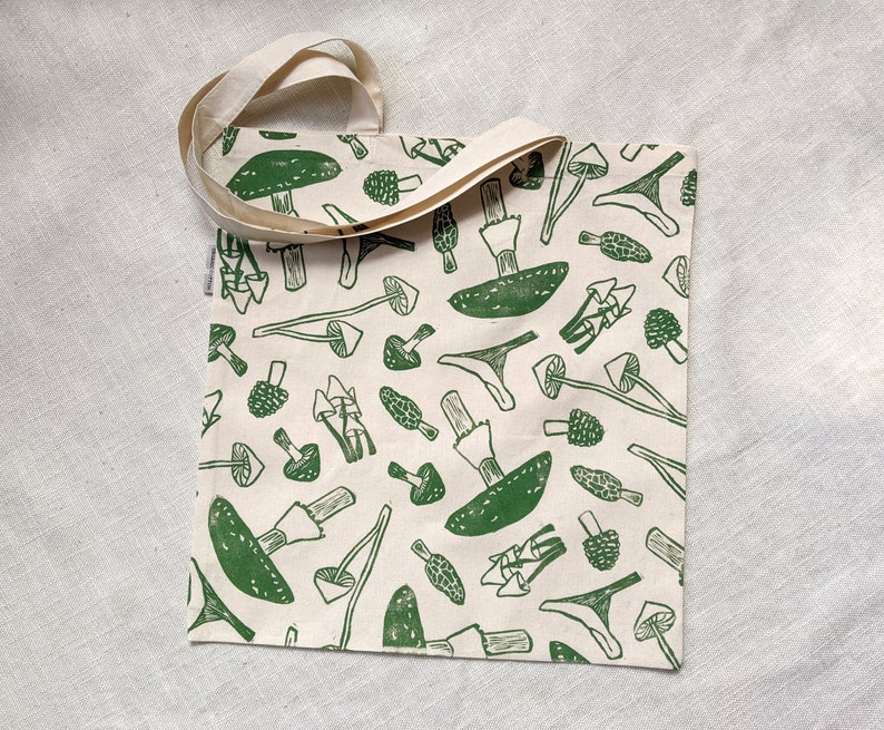 May include: A white canvas tote bag with green mushroom illustrations. The bag has two long straps.