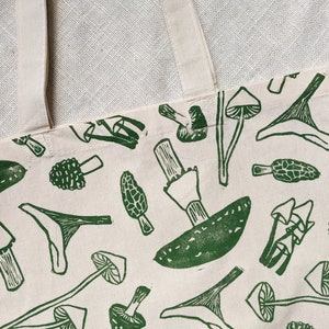 May include: A white canvas tote bag with green mushroom illustrations. The mushrooms are in various shapes and sizes, including some with stems and caps.