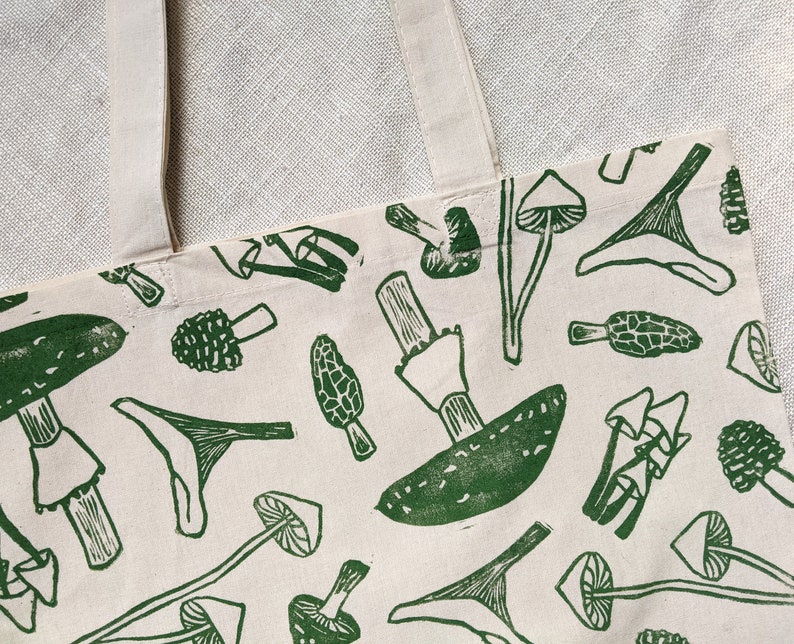 May include: A white canvas tote bag with green mushroom illustrations. The mushrooms are in various shapes and sizes, including some with stems and caps.