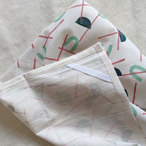 May include: Two white cotton tea towels with a geometric pattern of red lines, green and blue semi-circles, and dark blue circles. The towels are folded and the pattern is visible on both sides.