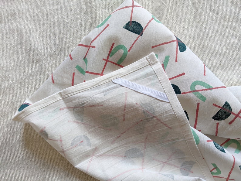 May include: Two white cotton tea towels with a geometric pattern of red lines, green and blue semi-circles, and dark blue circles. The towels are folded and the pattern is visible on both sides.