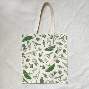 May include: A white tote bag with green mushroom illustrations. The mushrooms are drawn in a simple, blocky style and are repeated across the entire bag.