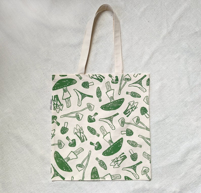 May include: A white tote bag with green mushroom illustrations. The mushrooms are drawn in a simple, blocky style and are repeated across the entire bag.